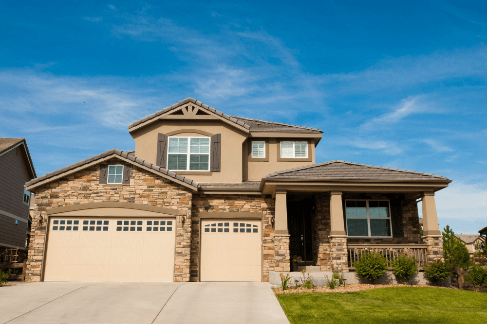 Two-story suburban home with a stone façade, beige three-section garage doors, and a covered front porch on a sunny day.